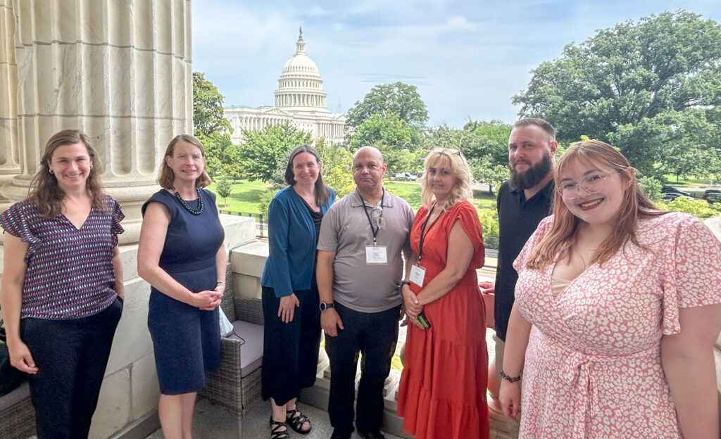 From left to right, Aliza Levine, Elizabeth O'Hara (Chief of Staff for Congressman Richard Neal), Sadie Simone, Dan Henderson, Cristina Welch, Kasey Mimitz, and Brianna Zimmerman stand together smiling on a balcony next to a large column and the Capitol Building in the distance behind them. 